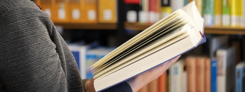 Eine Frau steht mit einem Buch in der Hand vor einem Bücherregal.