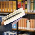 Eine Frau steht mit einem Buch in der Hand vor einem Bücherregal.